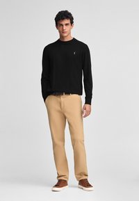 Maglione nero a maniche lunghe con un piccolo emblema, abbinato a pantaloni chinos beige e sneakers marroni. L'insieme presenta un design semplice e classico.