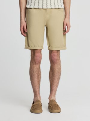 Mężczyzna ubrany w beżowe podwinięte szorty, brązowe buty wsuwane i biały teksturowany top bez rękawów na jednolitym tle.