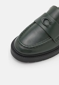 Mocassin en cuir vert foncé avec une semelle en caoutchouc noire et un détail décoratif en forme de fer à cheval en métal en relief sur la bride.