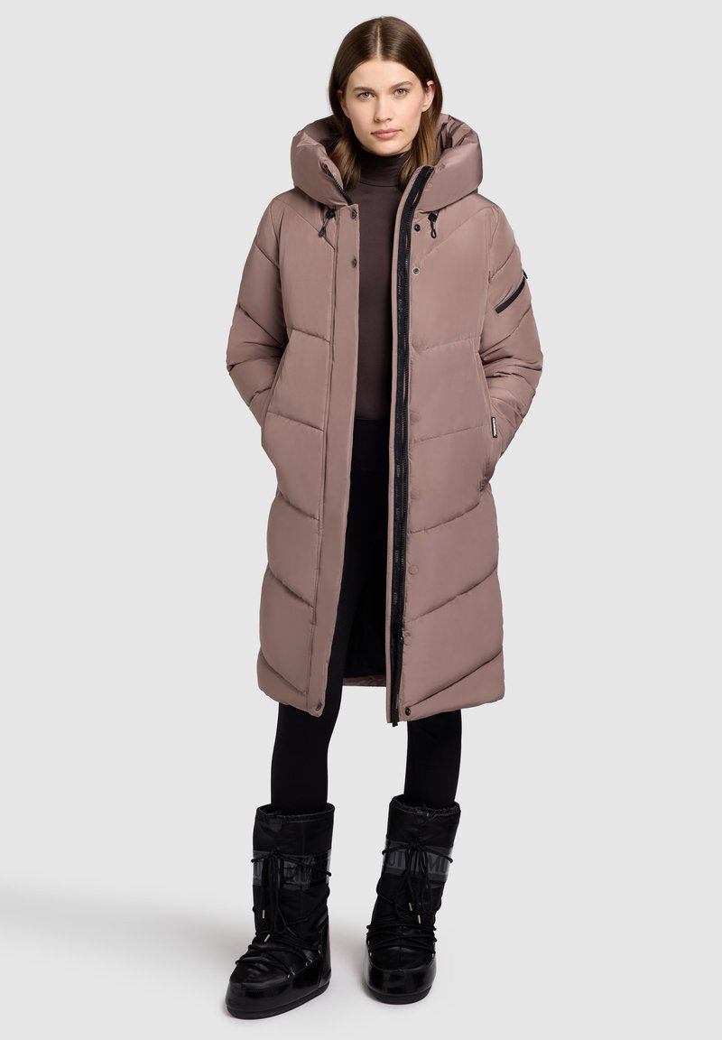 Long, padded coat in light taupe with a hood, zip closure, and side pockets. Paired with black, fur-lined winter boots.