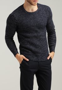 Granatowy sweter dzianinowy z teksturowanym wzorem, okrągłym dekoltem oraz ściągaczami przy rękawach i u dołu, w połączeniu z ciemnymi spodniami.