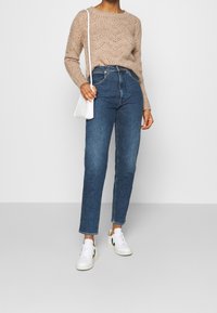 Suéter de punto beige con patrón de olas, jeans de mezclilla azul de cintura alta y zapatillas blancas con acentos negros. Bolso cruzado blanco.