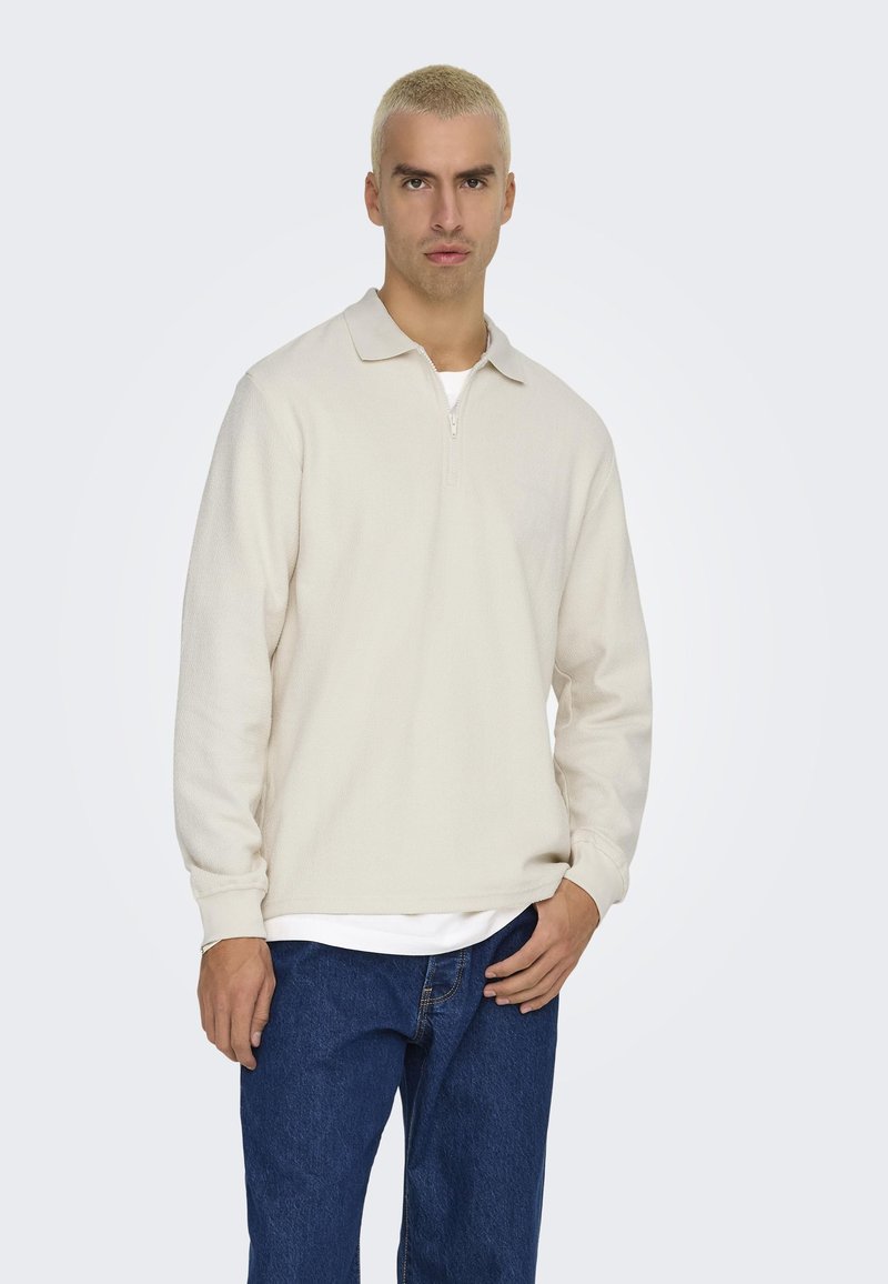 Pull crème à manches longues avec demi-fermeture éclair, col polo et tissu texturé, associé à un jean en denim bleu foncé.