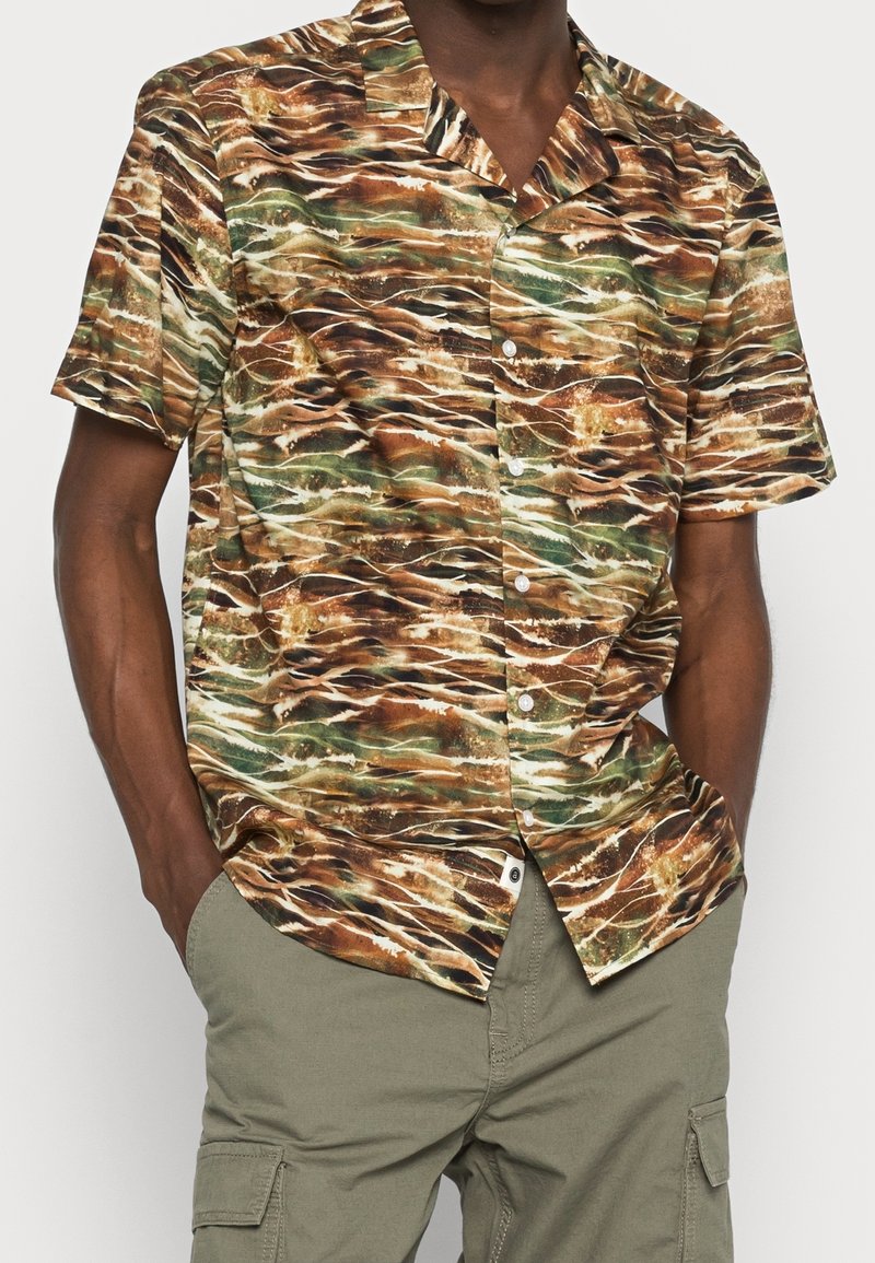 Homme portant une chemise à manches courtes boutonnée avec un motif abstrait marron et vert et un pantalon cargo vert olive, les mains dans les poches.