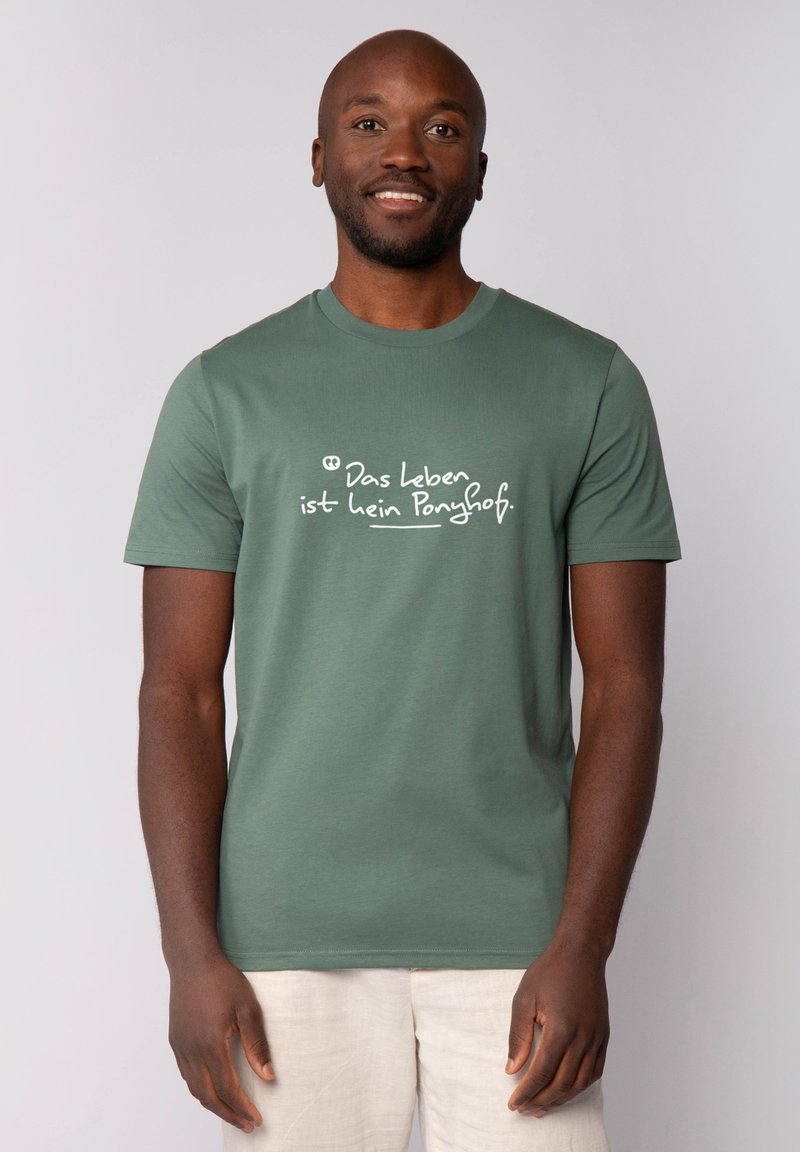Een glimlachende man in een groene T-shirt met witte Duitse tekst, beige broek, staande tegen een effen lichte achtergrond.