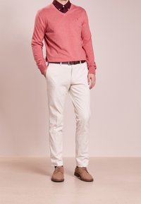 Homme portant un pull à col en V rose sur une chemise à col bordeaux, pantalon blanc avec une ceinture marron et des chaussures de ville en daim beige.