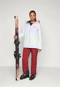 Naine valges suusajope ja punastes pükstes hoiab suuski vertikaalselt, jalas mustad suusasaapad, seistes ühtlase tausta ees.