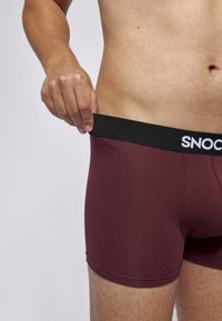 Bordó boxerské trenýrky s černým elastickým pasem, na kterém je bílé logo "SNOC". Materiál vypadá měkký, má hladký povrch a blízko přiléhající design.