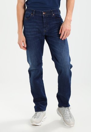 Uomo che indossa jeans blu scuro a gamba dritta, sneakers bianche e una camicia blu scuro, in piedi con le mani rilassate lungo i fianchi.