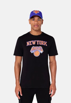 Uomo con t-shirt nera dei New York Knicks e cappellino da baseball blu dei Knicks, in piedi davanti a uno sfondo chiaro e uniforme.