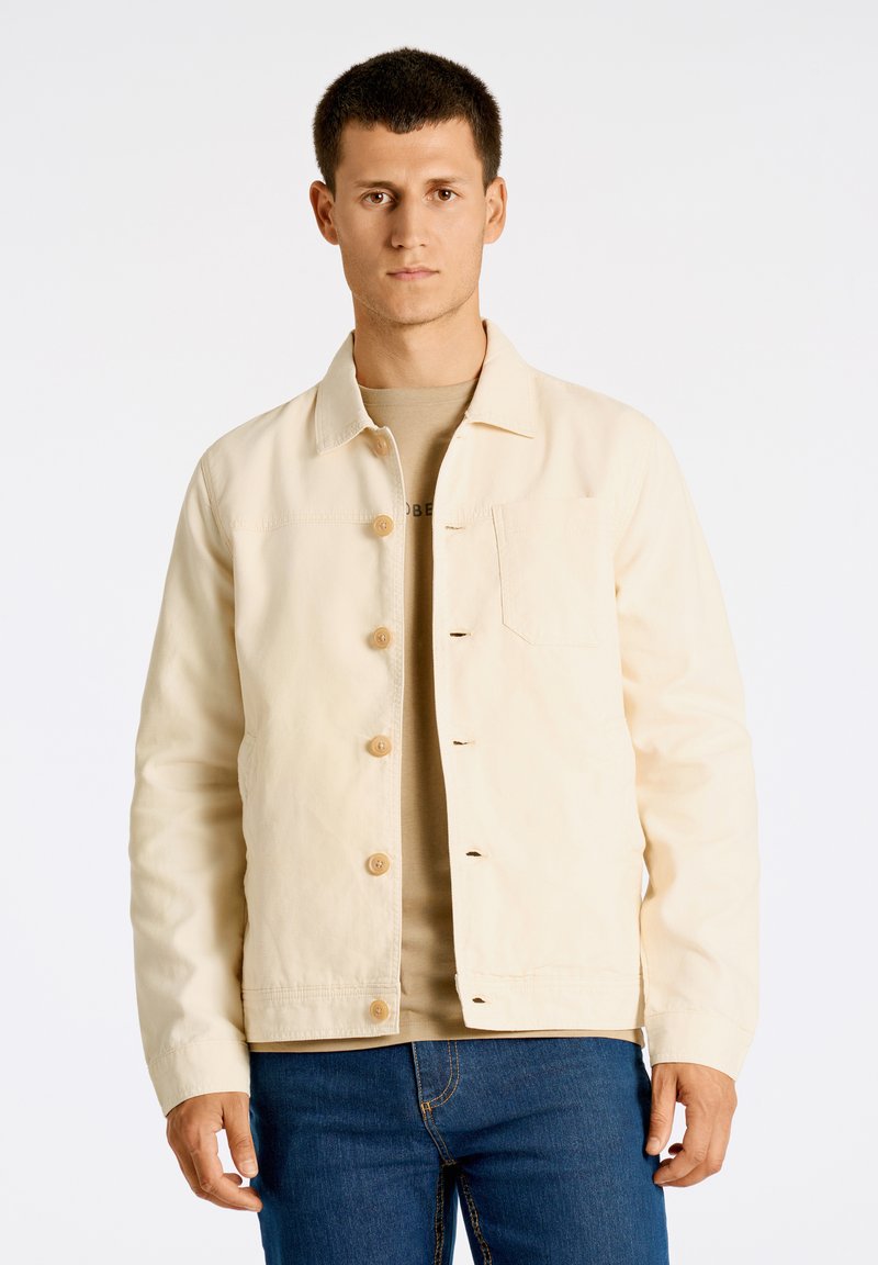 Beige bomullsjacka med krage, framknäppning och bröstfickor. Bärs över en ljusbrun skjorta och matchas med blå jeans.