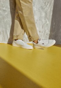 Osoba w beżowych spodniach i białych butach Tommy Hilfiger idąca po żółtej podłodze na tle szarej, teksturowanej ściany.