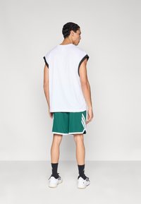 Weißes ärmelloses Shirt mit schwarzen Akzenten, grüne Shorts mit weißen Streifen, schwarze Socken und weiße Turnschuhe, von hinten gesehen.