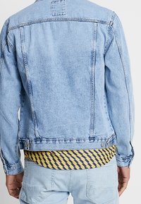 Homme portant une veste en denim bleu clair sur une chemise jaune avec un motif d'éclairs bleus, vu de dos.