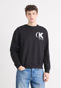 Sudadera neagră cu guler rotund și mâneci lungi, având un logo alb "CK" și textul "Calvin Klein" pe pieptul stâng. Pur tată cu blugi albaștri.