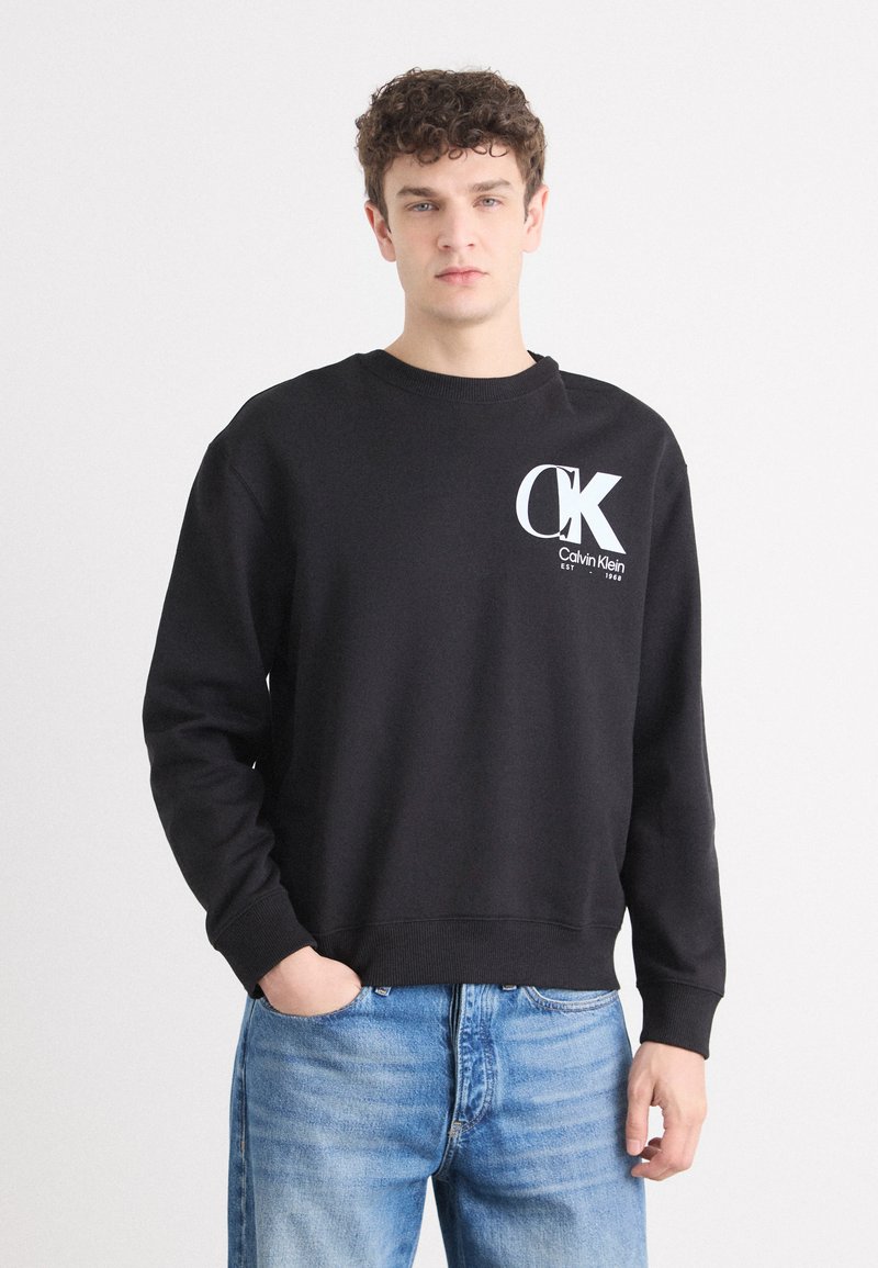 Sudadera neagră cu guler rotund și mâneci lungi, având un logo alb "CK" și textul "Calvin Klein" pe pieptul stâng. Pur tată cu blugi albaștri.