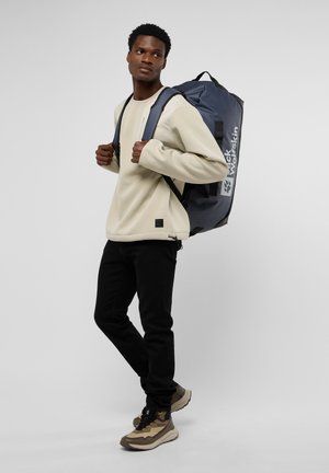 Mochila en azul oscuro con detalles en negro, correas ajustables y logo. Modelo vistiendo un suéter beige y pantalones negros, de pie de manera casual.