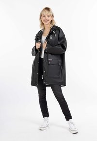 Impermeable negro con capucha, botones a presión, bolsillos frontales y un corte relajado, llevado sobre una blusa blanca y jeans negros.