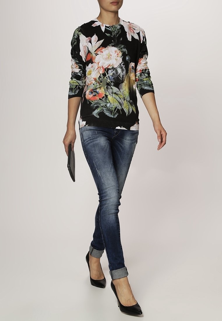 Femme marchant portant une chemise à manches longues à fleurs, un jean bleu retroussé, des talons hauts noirs et tenant une pochette noire dans une main.
