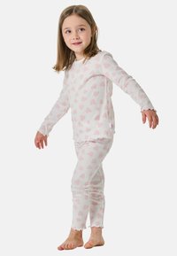 Junges Mädchen in weißen, gerippten Pyjamas mit rosa Herzmustern, barfuß stehend mit leicht ausgestreckten Armen vor einem schlichten Hintergrund.
