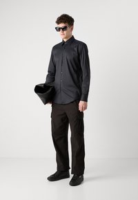 Camisa negra de botones con un acabado brillante, combinada con unos pantalones cargo negros holgados. Los accesorios incluyen gafas de sol negras de gran tamaño y un bolso negro.