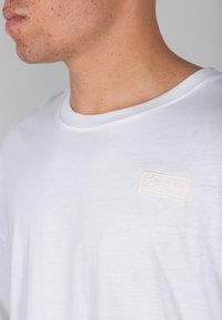 T-shirt bianco in cotone con collo rotondo e un'etichetta cucita color crema sul lato sinistro del petto che riporta "ALPHA INDUSTRIES". Texture liscia.