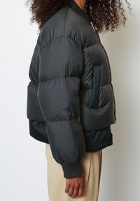 Black padded bomber jacket with a ribbed cuff and collar, featuring a side pocket and smooth texture. Overlaid on light beige pants.