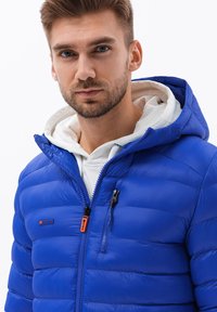 Giacca a vento blu con cappuccio, caratterizzata da una chiusura con zip, cuciture orizzontali e un accento arancione sul lato sinistro. Fodera interna bianca.