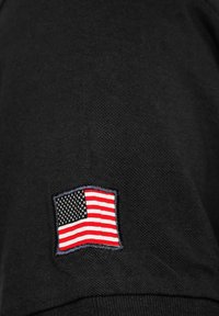 Schwarzer Stoff mit einer strukturierten Oberfläche, der an der Seite mit einem kleinen, gestickten amerikanischen Flaggen-Patch in Rot, Weiß und Blau versehen ist.