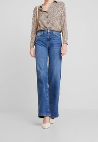 Blusa de estampado de leopardo con mangas largas, cierre de botones en el frente, combinada con unos jeans de pierna ancha y cintura alta de color azul. Tacones de color claro completan el look.
