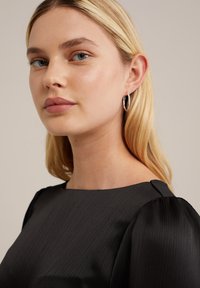 Abito in tessuto nero con texture, scollatura a quadrato e maniche a sbuffo. Il modello indossa orecchini a cerchio in argento. I suoi capelli morbidi e naturali le cadono sulla spalla.