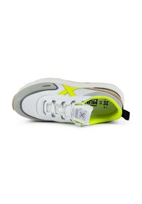 Chaussure de sport blanche avec des accents gris, des détails en néon jaune et une semelle texturée. Présente des lacets, un patch de logo et un talon renforcé.