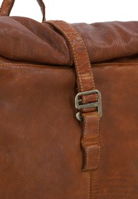 Sac en cuir marron avec une surface texturée. Il dispose d'un haut enroulé et d'un détail de sangle avec boucle en métal pour la fermeture. Les bords cousus sont visibles.