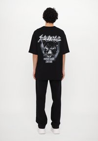 Juodas per didelis t-shirt'as, vaizduojantis panterą ir tekstą "Sauvage" bei "Versace Jeans Couture" baltos ir sidabrinės spalvos. Atskleistas su juodomis kelnėmis.