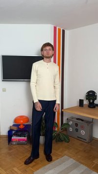 Krém színű, hosszú ujjú póló, henley nyakkal és három gombbal, sötétkék nadrággal és barna cipőkkel párosítva, egy díszített szobában állva.