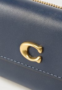 Πορτοφόλι από δέρμα σε ναυτικό μπλε χρώμα με υφή στην επιφάνεια, εξαρτήματα σε χρυσό χρώμα και αντίθετη ραφή σε λευκό κατά μήκος της άκρης. Κλείσιμο με φερμουάρ.