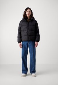 Sort pufferjakke med hætte, quiltet design, frontlynlås, elastiske manchetter. Kombineret med løse blå jeans og lyse sko.