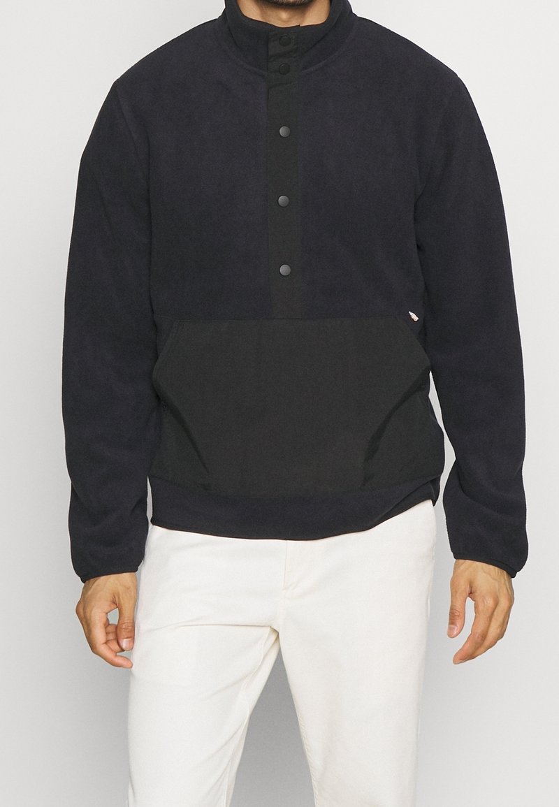 Homme portant un pull polaire noir avec des boutons-pression et une poche avant, associé à un pantalon blanc sur un fond uni.