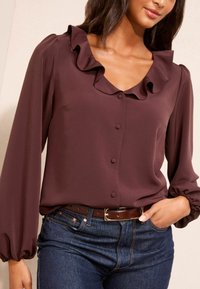 Maroon blūze ar volānu apkakli, garām pūstajām piedurknēm un pogājamu priekšu. Saskaņota ar tumšas džinsu biksēm un brūnu jostu.
