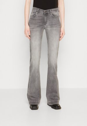 Person iført højttaljede, grå udvaskede flare-jeans og sorte sko med firkantet tå, stående mod en ensfarvet hvid baggrund.