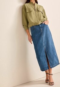 Mujer vestida con blusa verde oliva con cremallera, falda midi de mezclilla azul con abertura frontal y tacones negros con tira al tobillo, de pie contra una pared clara.