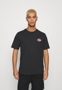 Camiseta de algodón negra con cuello redondo, que presenta un pequeño logotipo gráfico rojo en el lado izquierdo del pecho. Mangas cortas; ajuste casual.