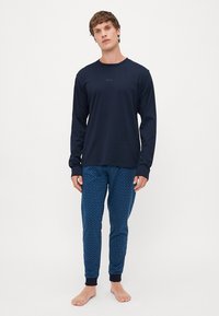 Camisa de manga longa da marinha com um logotipo discreto, combinada com calças de pijama azuis estampadas com punhos canelados. O modelo está descalço.