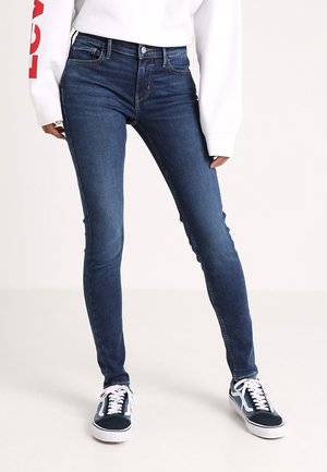 Personne portant un jean skinny bleu foncé, un sweat-shirt blanc avec un détail rouge sur la manche, et des baskets bleu marine avec des lacets blancs sur fond uni.
