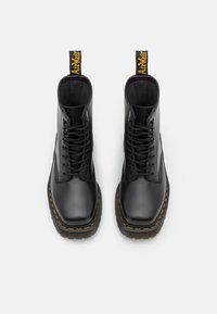 Stivali alla caviglia in pelle nera con punta rotonda, chiusura con lacci, cuciture gialle e un linguetta con la scritta "AirWair". Design della suola piatta.