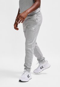 Graue Jogginghose aus weichem Material, mit Slim-Fit-Schnitt, seitlichen Taschen und einem kleinen weißen Logo am linken Oberschenkel. Getragen mit weißen Sneakern.
