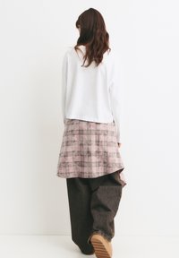 Weißes Langarmoberteil, das mit einem rosa karierten Hemd geschichtet ist, mit asymmetrischem Saum. Locker sitzende dunkle Jeans mit einer hellbraunen Sohle.