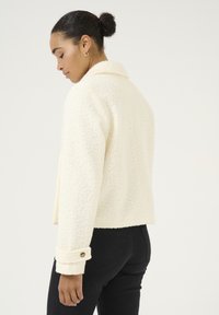Veste en polaire texturée couleur crème, avec un col, un design court et des poignets boutonnés, associée à un pantalon noir ajusté.