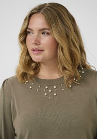 Vrouw met schouderlang golvend blond haar, gekleed in een olijfgroen sweater versierd met verspreide parelparels bij de halslijn.