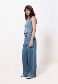 Blauer Jeansoverall mit angepasstem Oberteil, weit geschnittenen Hosenbeinen, durchgehender Knopfleiste und vertikalen Nähdetails. Glatte Textur, ärmelloses Design.